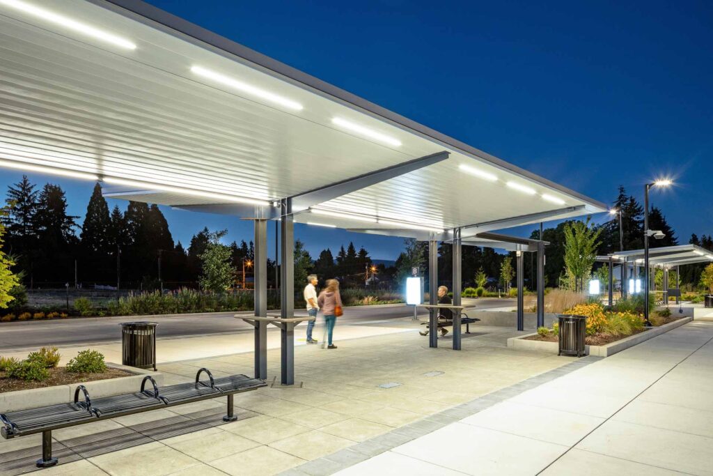 A modern outdoor transit station at dusk with people standing under well-lit shelters, benches, trash bins, and landscaped surroundings.