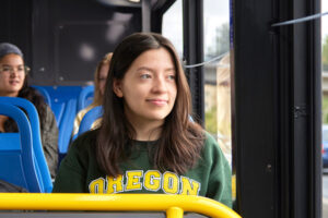 A young woman wearing an "Oregon" sweatshirt sits on a bus, looking out the window, with other passengers seated nearby.