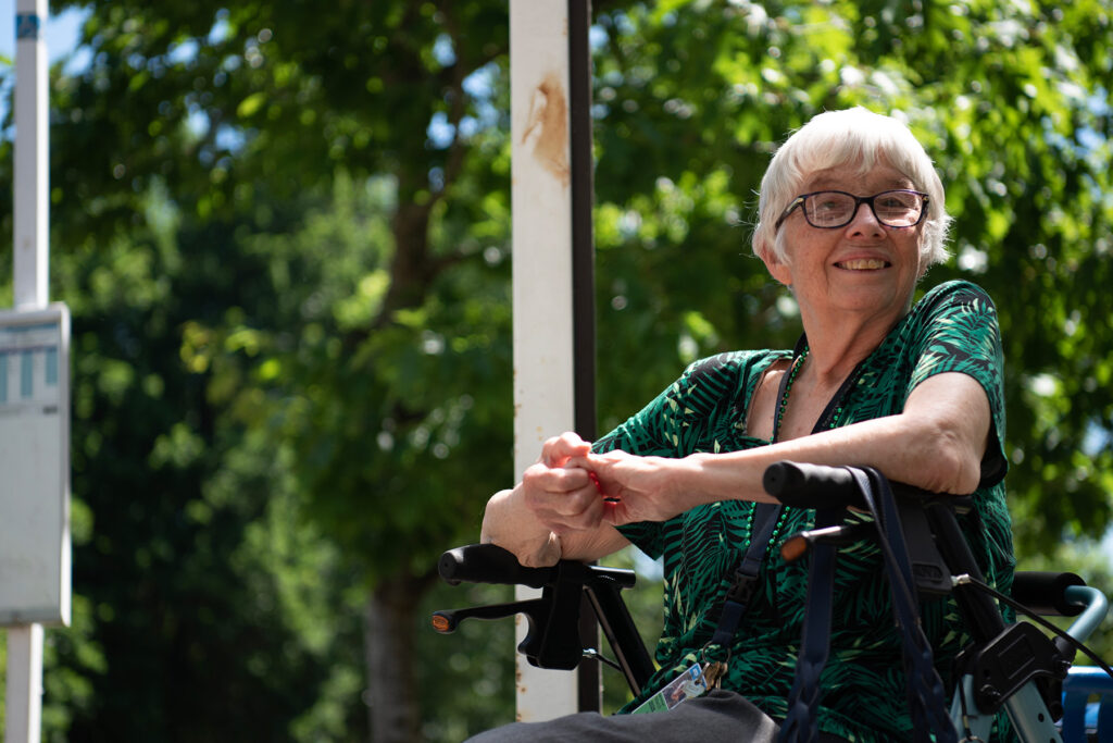 An elderly woman with short white hair and glasses sits in a wheelchair outdoors, smiling. Trees and greenery are visible in the background.