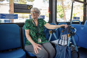 An older woman with glasses sits on a bus, smiling, with a walker and handbag next to her. Signs for mobility device securement and wheelchair seating are visible.