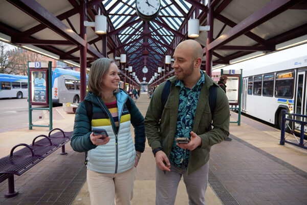 Two people with backpacks walk and talk at a bus station, holding phones. Buses and a clock are visible in the background under a glass roof.
