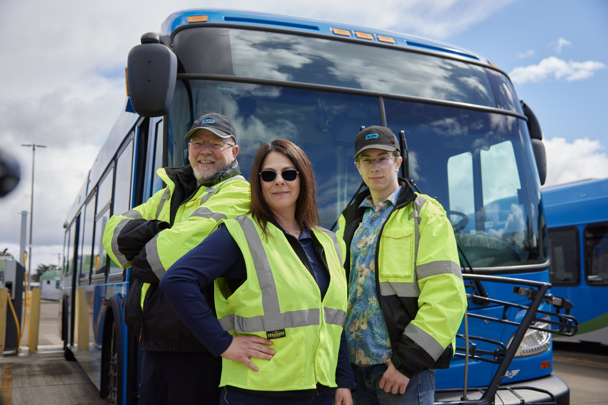 Three people in yellow safety vests stand confidently in front of a blue city bus, posing for the camera at a bus depot.