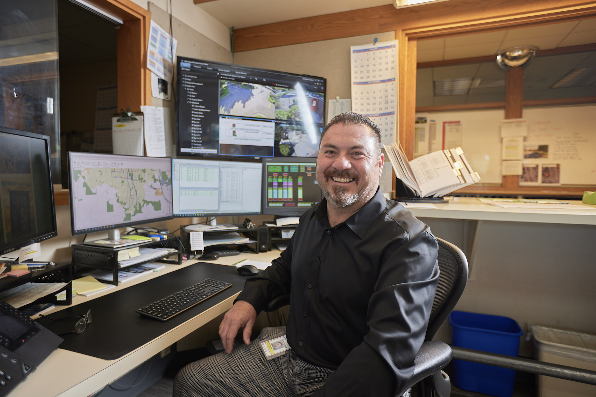 A man in a black shirt sits and smiles at a workstation with multiple monitors displaying maps, graphs, and data in an office setting.