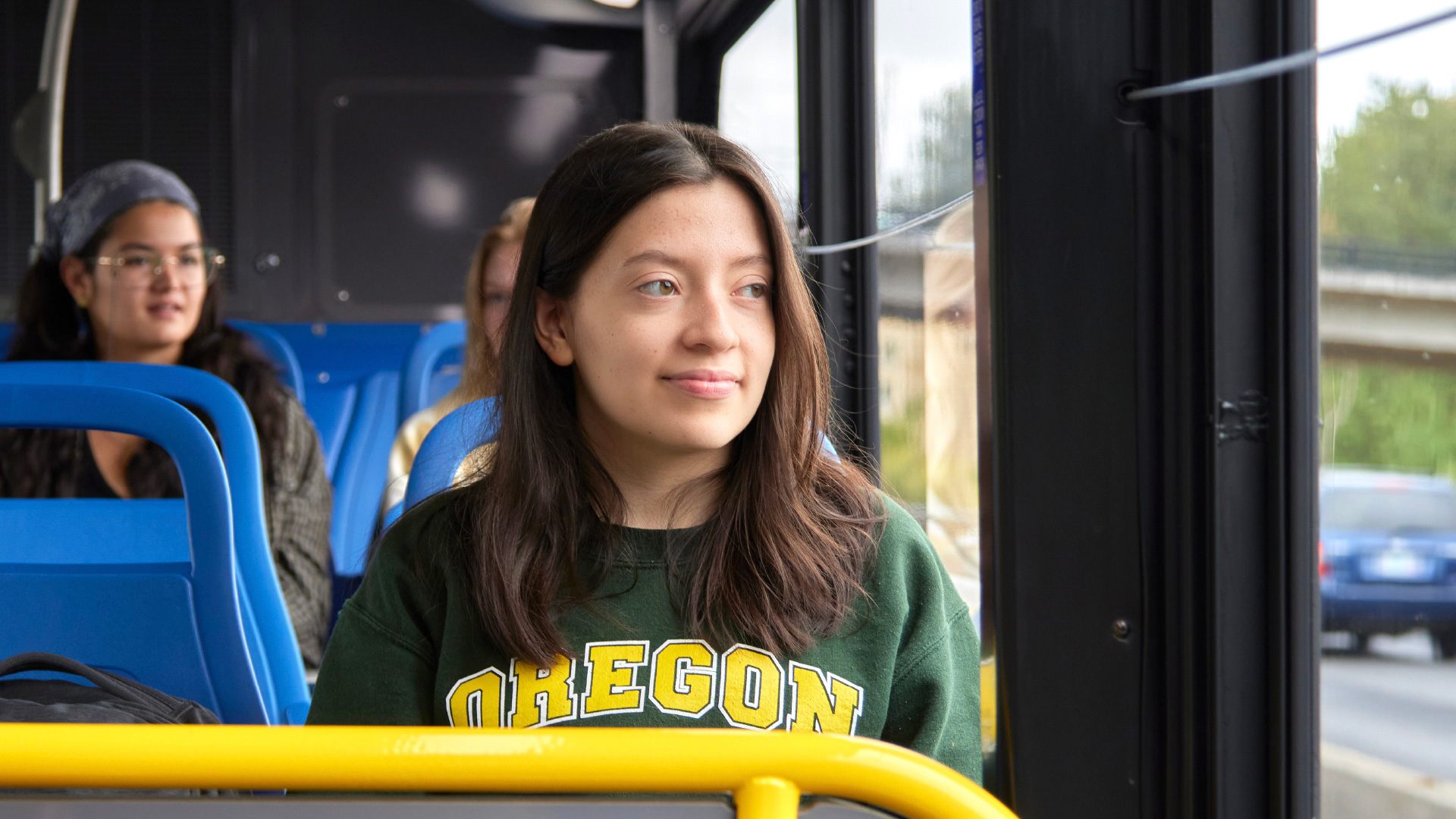 A young woman wearing an "Oregon" sweatshirt sits on a bus, looking out the window. Other passengers are seated behind her.