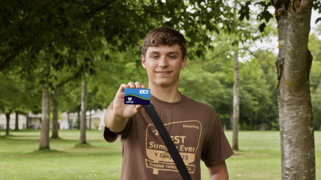 A young person standing outdoors holding a blue Umo card toward the camera, wearing a brown camp t-shirt and a crossbody bag. Trees and grass are visible in the background.