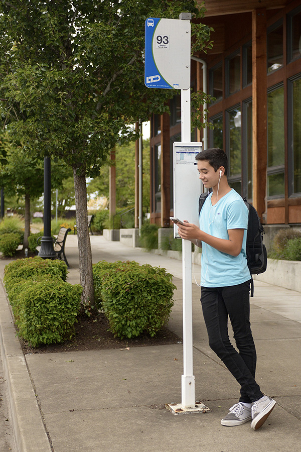 Teenager with a backpack stands at a bus stop, listening to earphones and using a phone. A bus stop sign and a building are visible in the background.