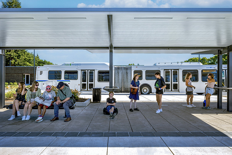 People wait at an outdoor bus stop under a shelter, with a white hybrid bus parked in the background on a sunny day.