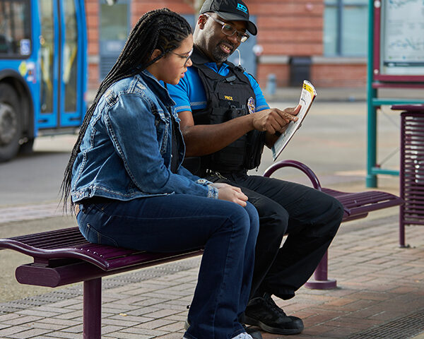 A security officer sits on a bench at a bus stop, showing a pamphlet to a woman seated next to him; a blue bus and map are visible in the background.