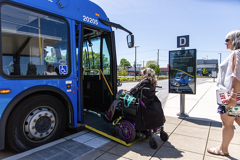 A person in a wheelchair boards a blue city bus using a ramp while another person waits nearby at a bus stop.