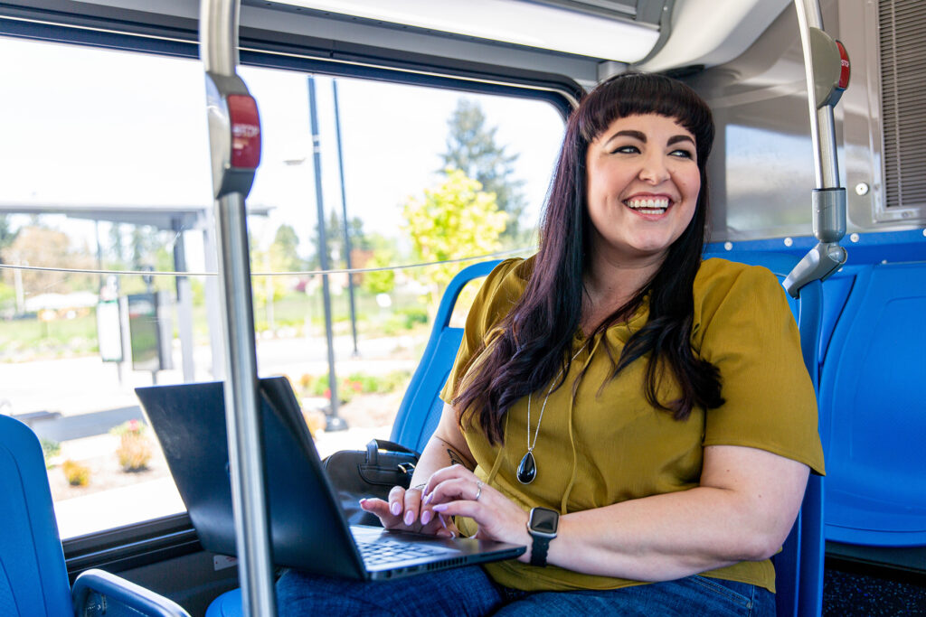 Woman with long dark hair sits on a blue bus seat, smiling and typing on a laptop, with a window showing greenery in the background.