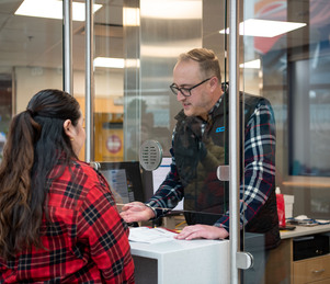 A man and a woman are speaking across a glass partition at a service counter, with paperwork and office supplies on the counter.