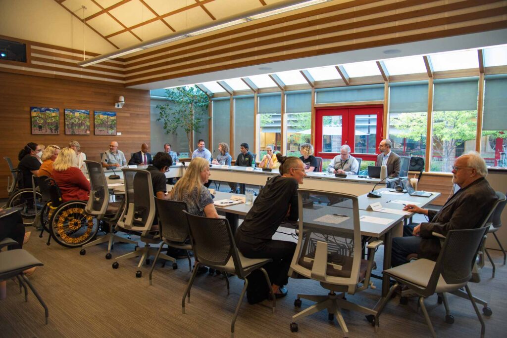 A group of people sit around a large conference table in a modern meeting room with large windows and natural light.