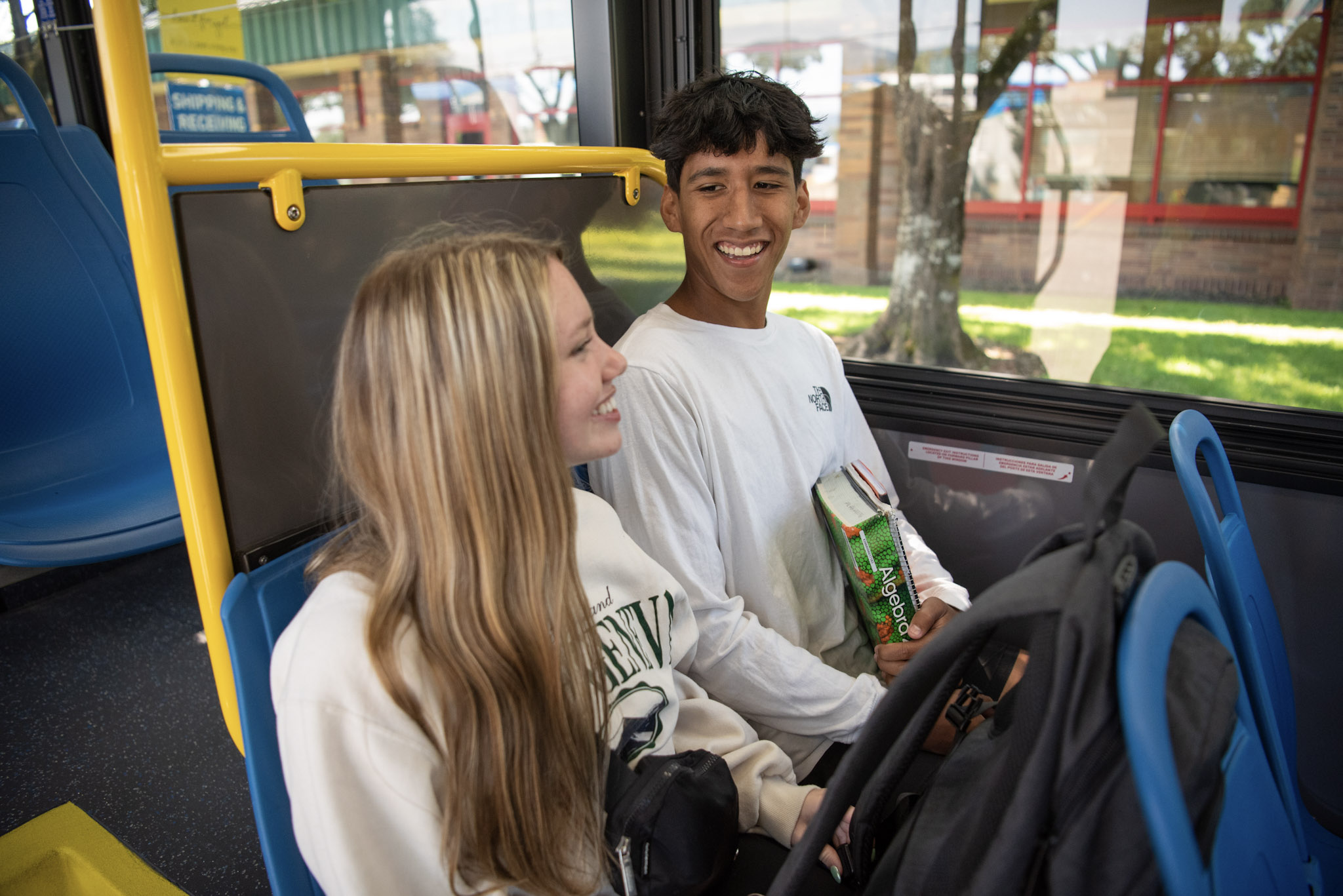 Two teenagers sit and talk on a bus; one holds a package of snacks while backpacks rest on their laps.
