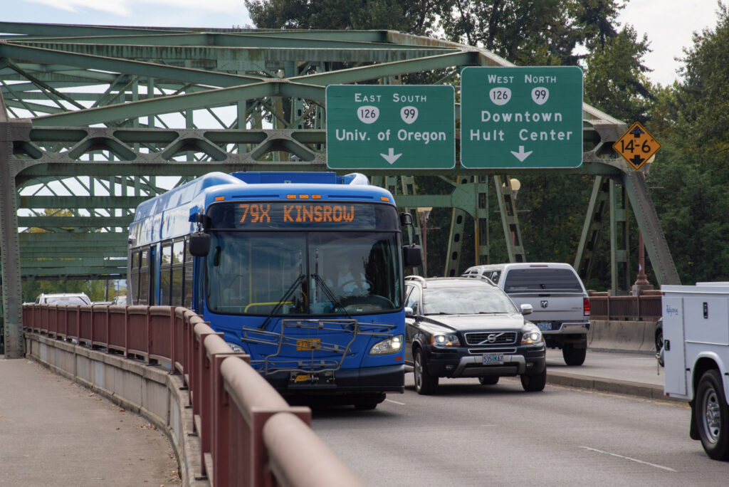 A blue city bus displaying "79x Kinsrow" drives across a bridge alongside cars, with green directional signs for the University of Oregon and Downtown in the background.