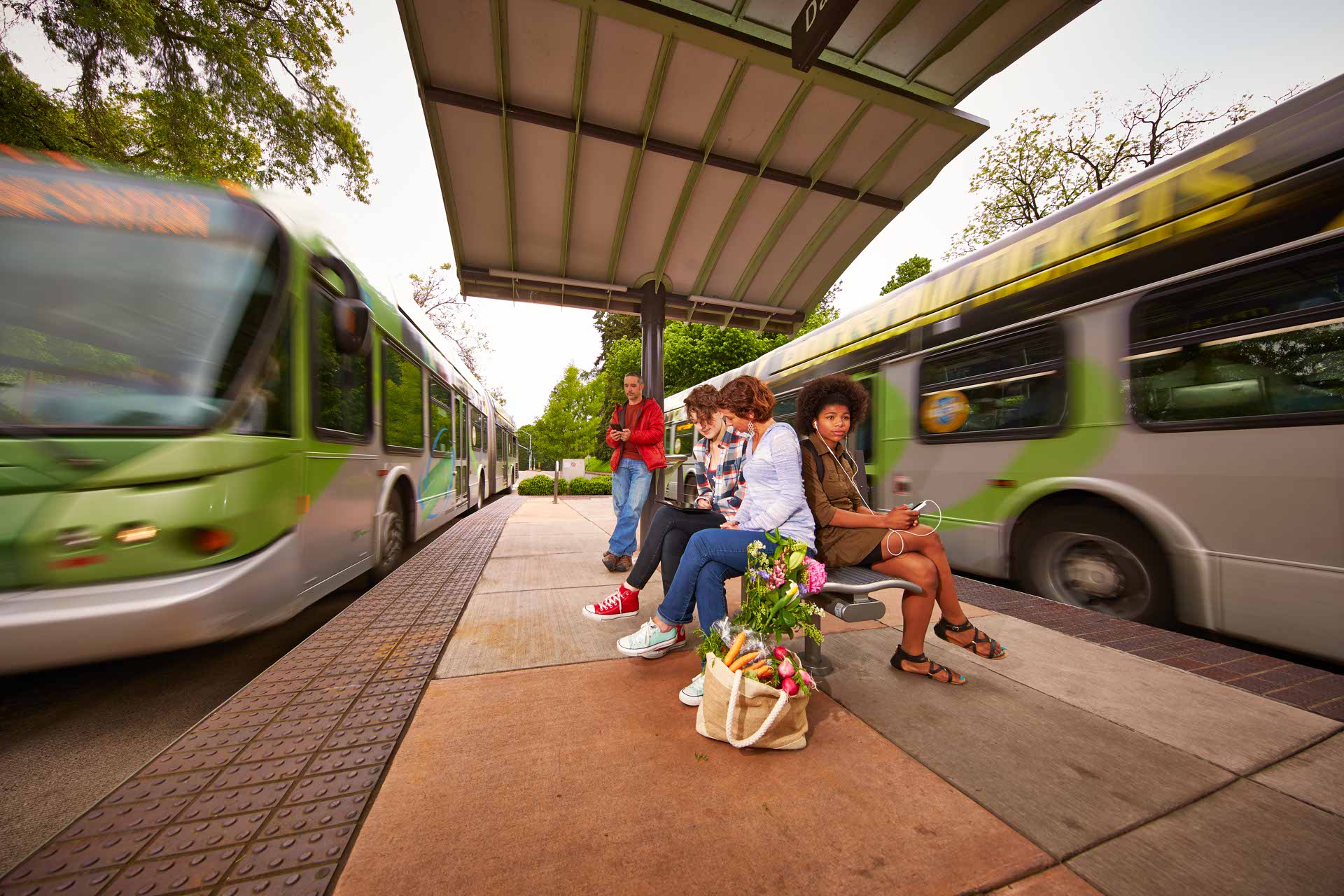 Three people sit on a bench at a bus stop, one holding flowers, while two buses move past on either side and another person stands nearby.