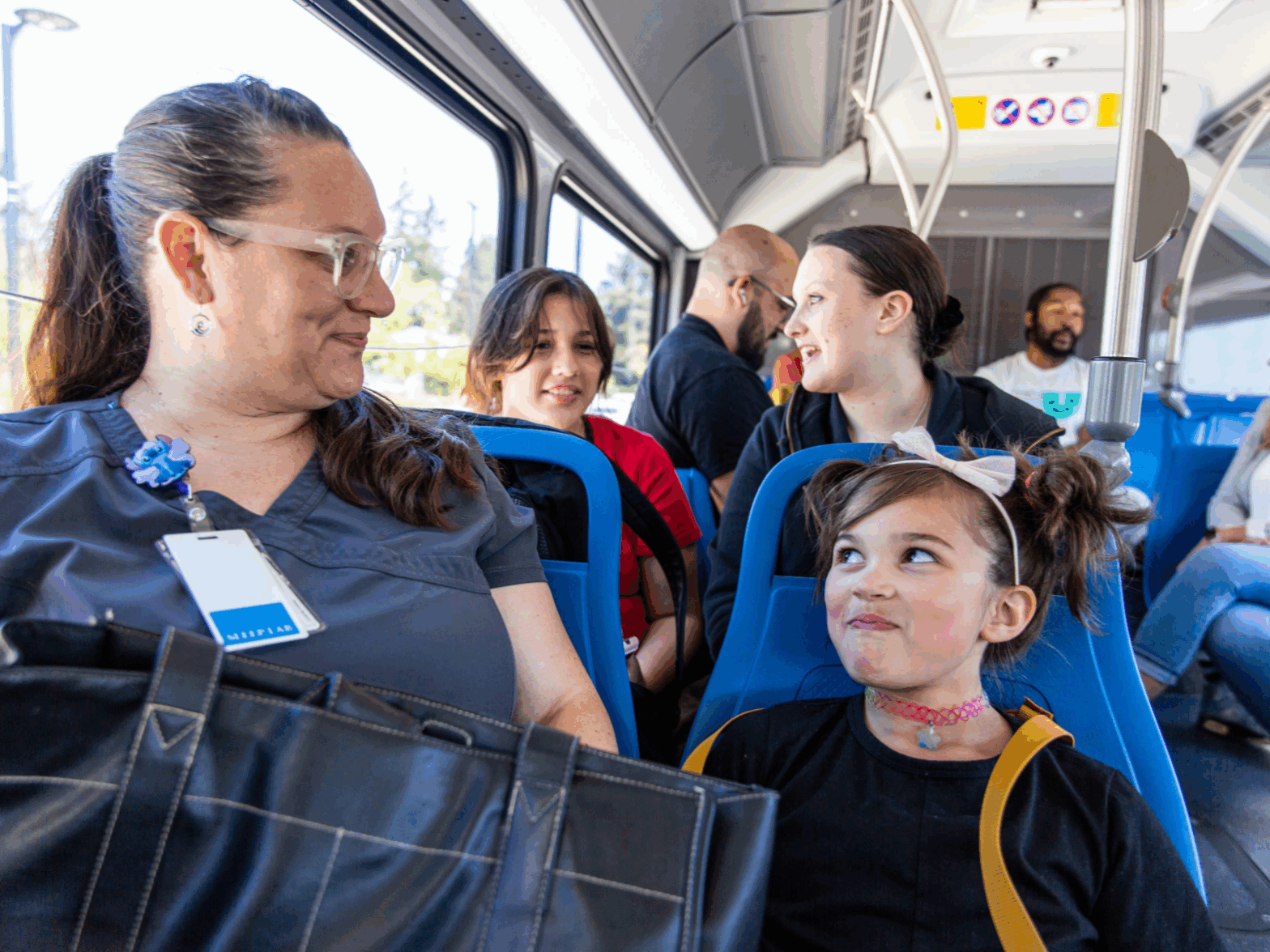 A woman with a badge and a young girl sit together on a bus, surrounded by other passengers, all engaged in conversation.