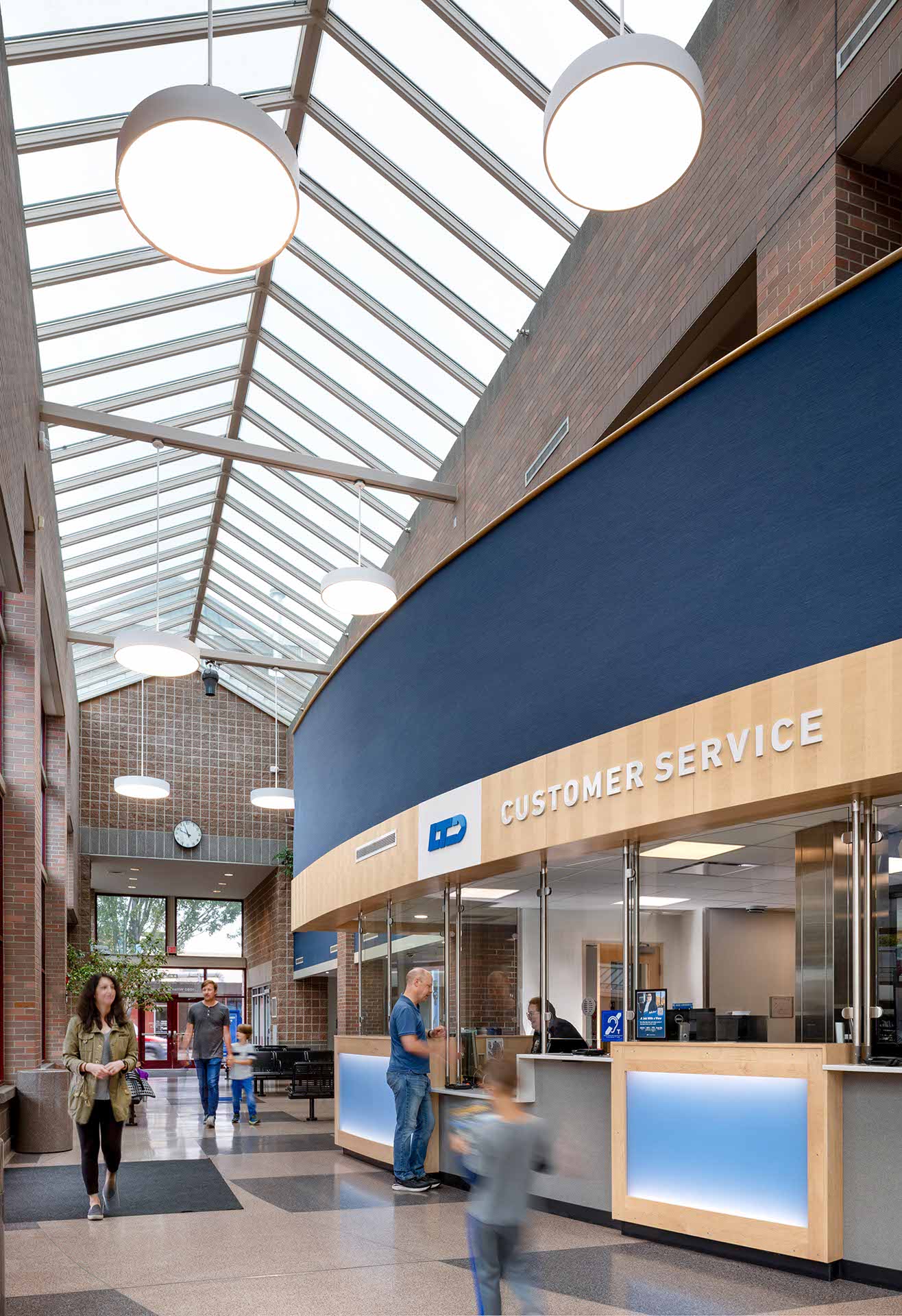 A modern customer service area with large windows, blue accents, and people interacting at the counter under a glass ceiling.