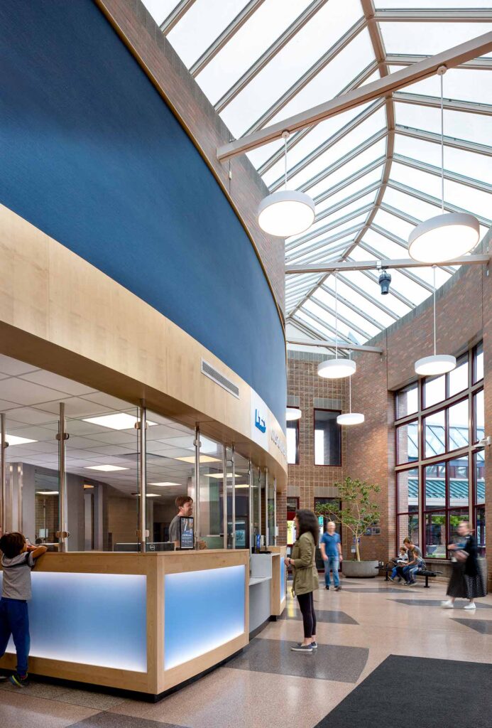 People stand and walk in a spacious, modern lobby with large windows, a glass ceiling, and a reception desk under blue accent walls.