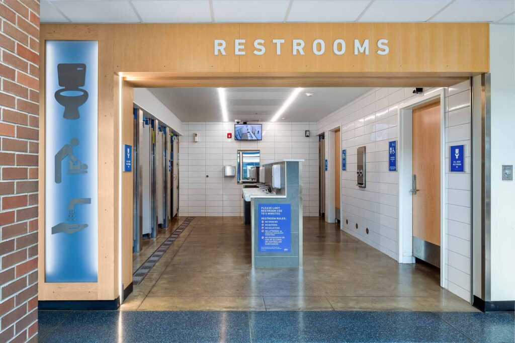 Public restroom entrance with clear signage, individual stalls, sinks, and hygiene icons displayed on the left wall.
