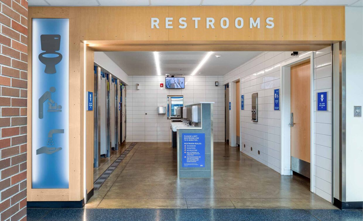 Public restroom entrance with clear signage, individual stalls, sinks, and hygiene icons displayed on the left wall.