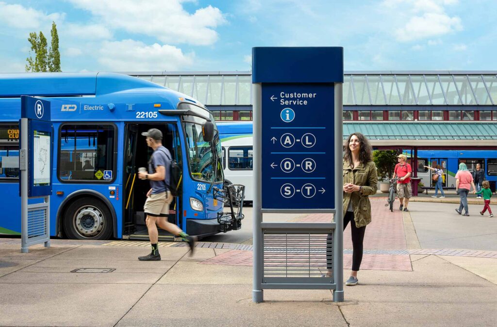 A blue electric bus is parked at a transit station. A man walks past, and a woman stands near a customer service sign with route information. Several people are in the background.