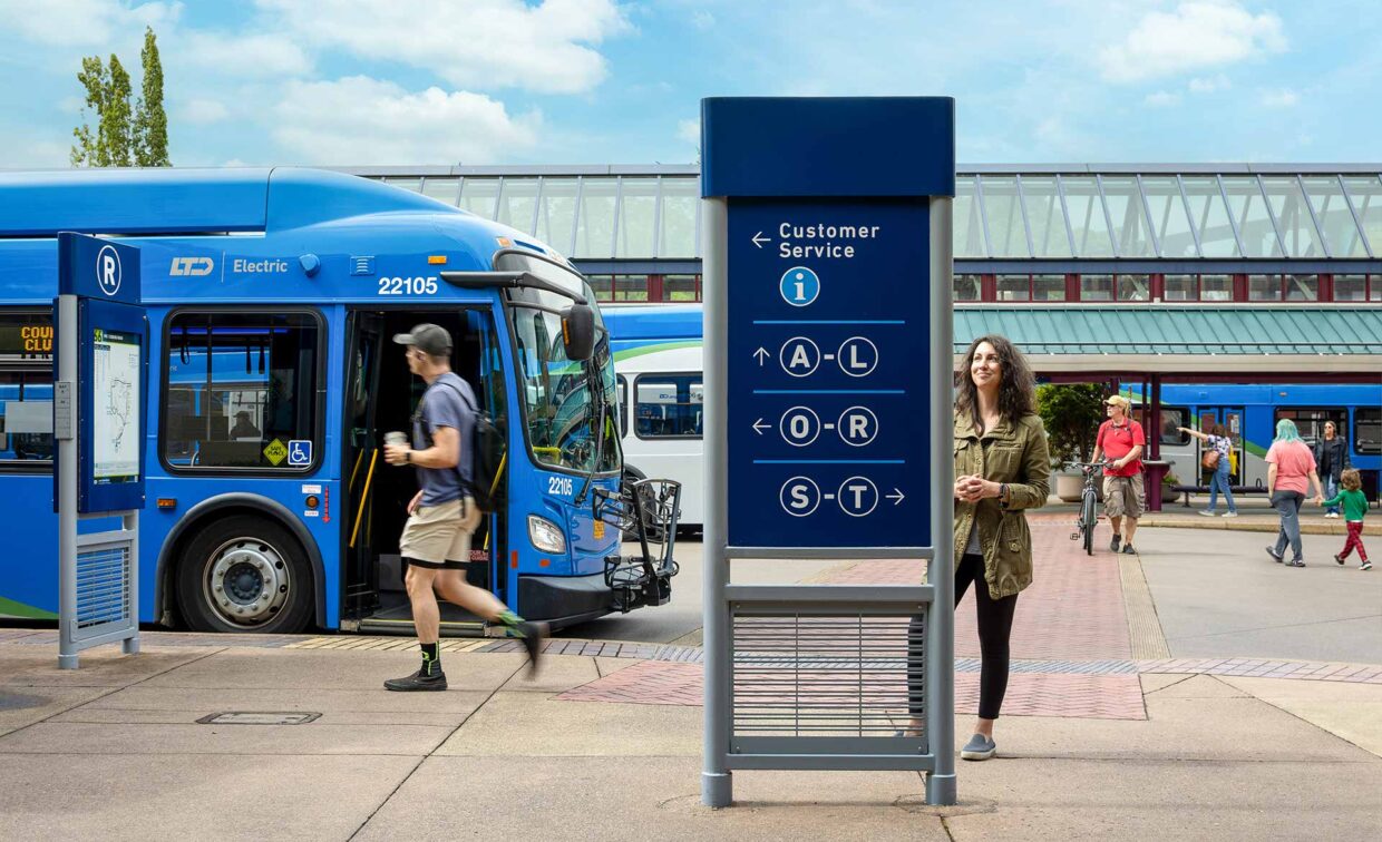 A blue electric bus is parked at a transit station. A man walks past, and a woman stands near a customer service sign with route information. Several people are in the background.