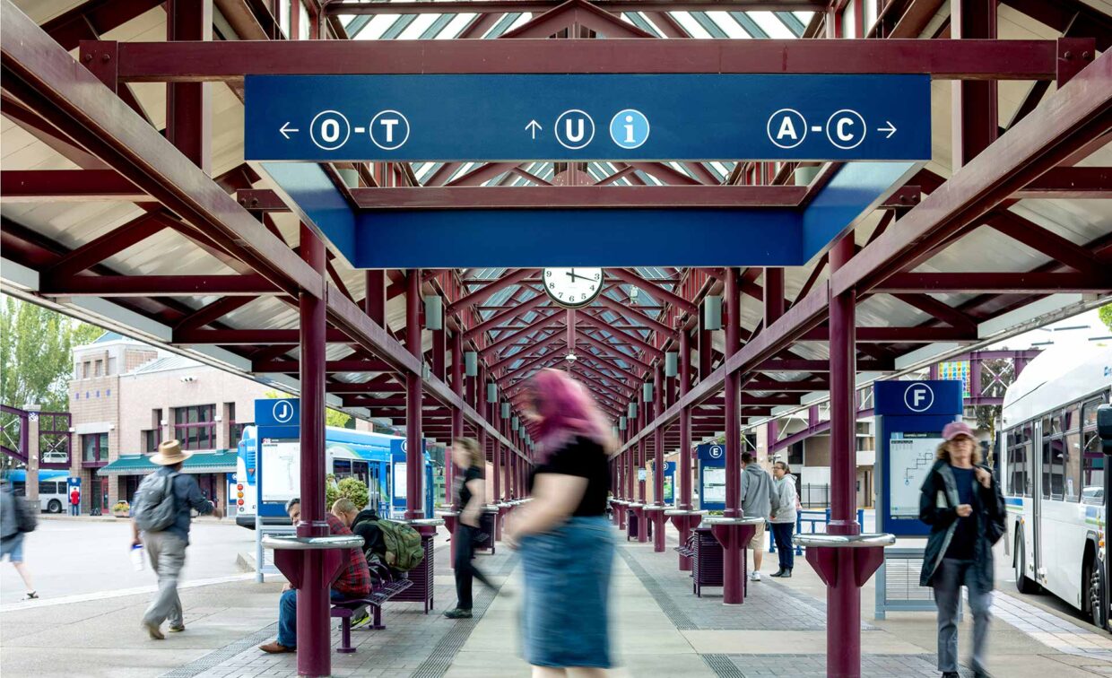 People walking and waiting at a covered outdoor transit station with blue directional signs and a clock overhead.