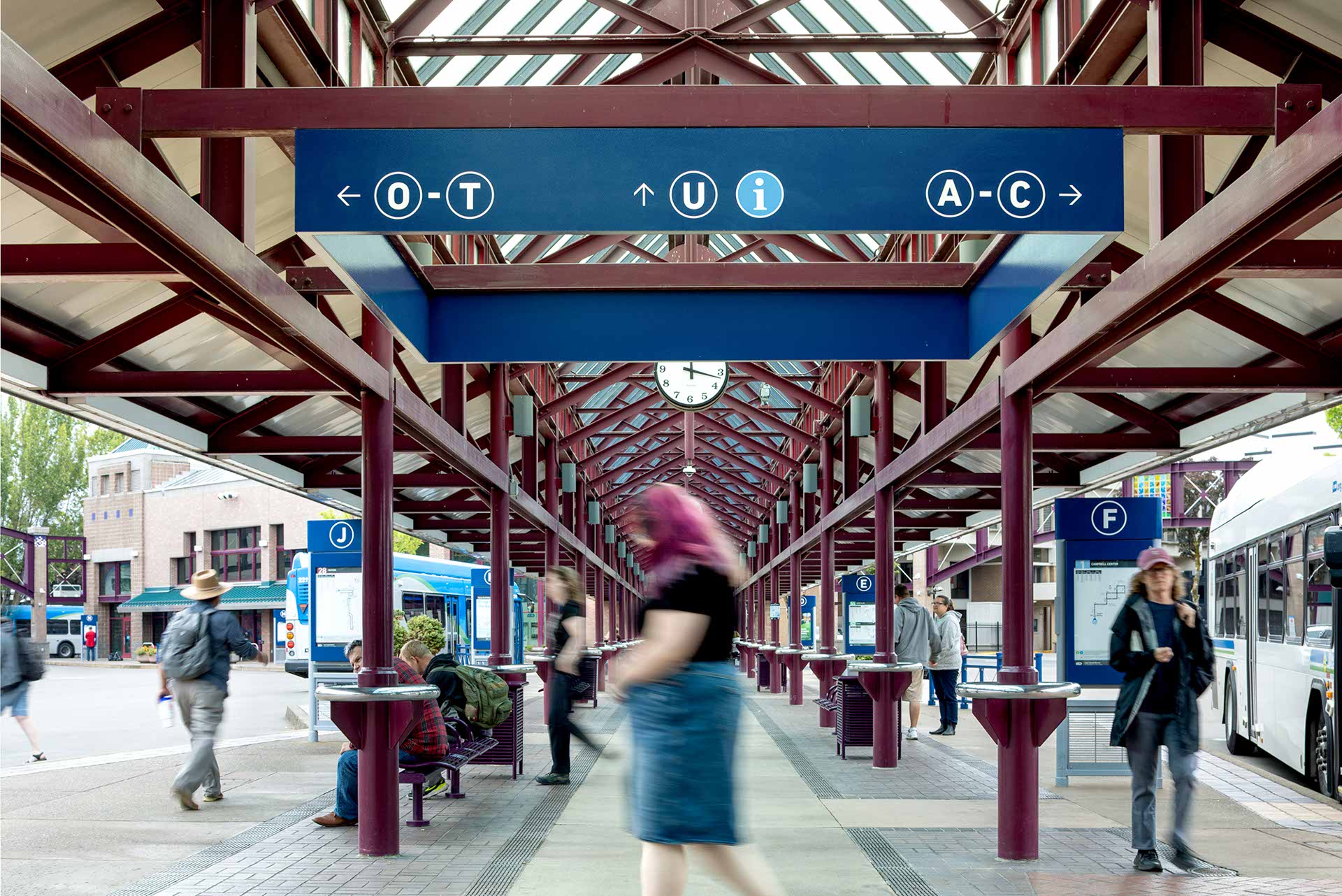 People walking and waiting at a covered outdoor transit station with blue directional signs and a clock overhead.