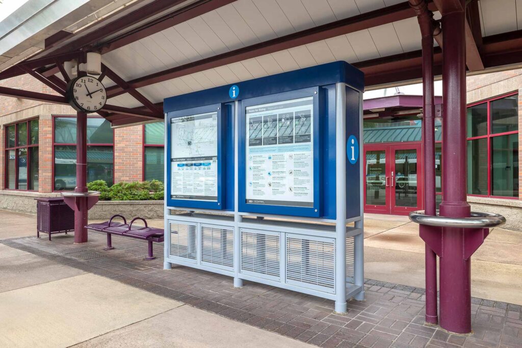 A transit information board with maps and schedules stands under a covered area near a wall clock, outdoor seating, and a building with glass doors and windows.