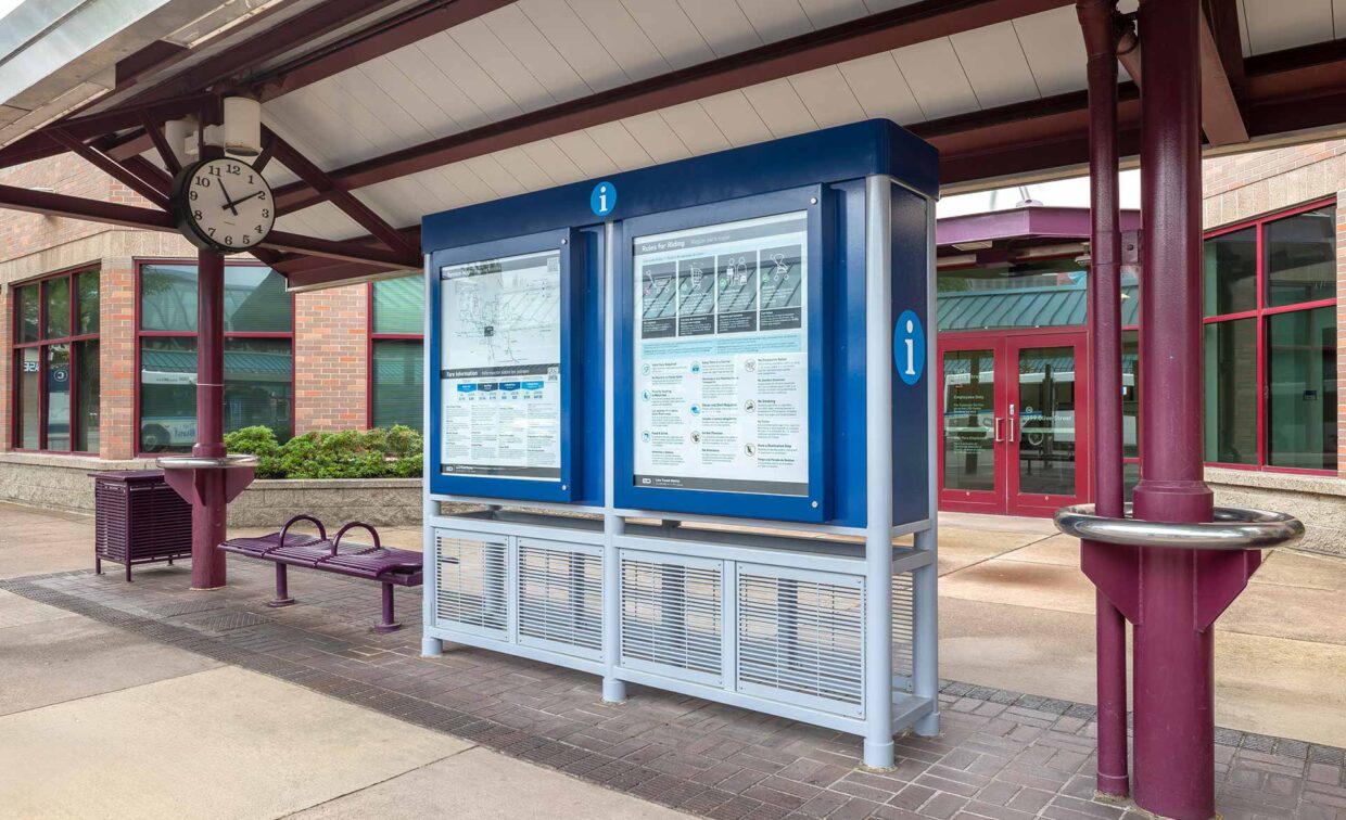 A transit information board with maps and schedules stands under a covered area near a wall clock, outdoor seating, and a building with glass doors and windows.