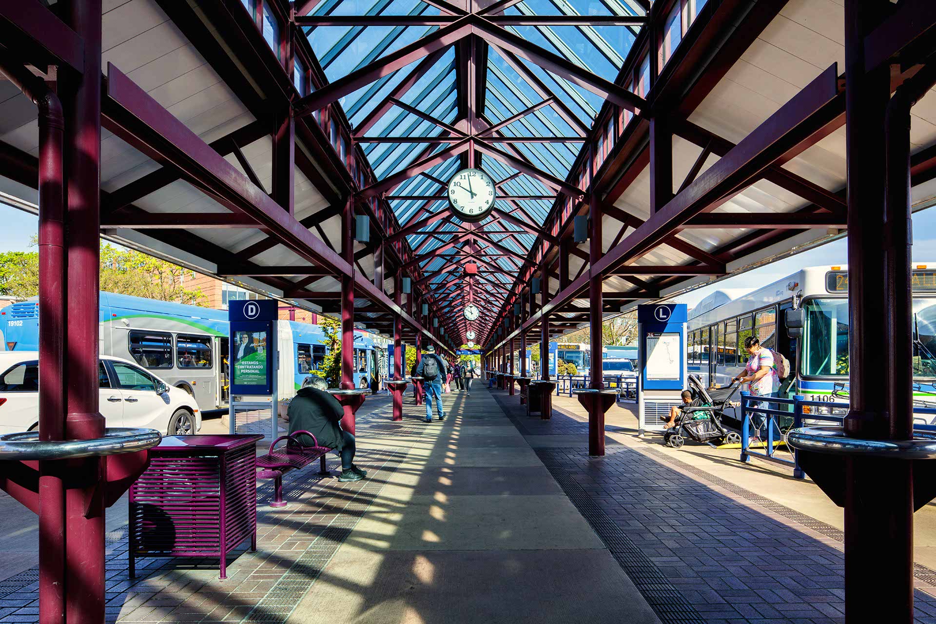 Covered outdoor bus terminal with a glass roof, multiple buses parked on both sides, benches, digital displays, and a clock suspended from the ceiling.
