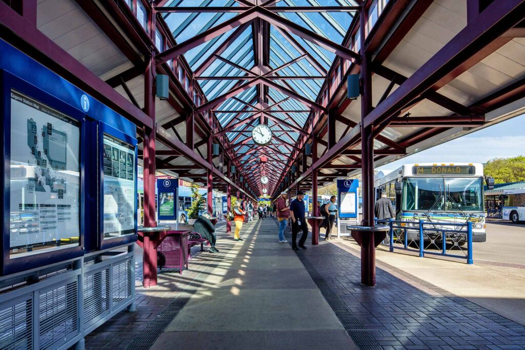 Covered outdoor bus terminal with a glass roof, passengers waiting, buses parked on the right, and route maps displayed on the left. A large clock hangs above the walkway.