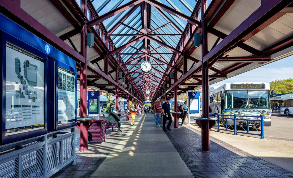 Covered outdoor bus terminal with a glass roof, passengers waiting, buses parked on the right, and route maps displayed on the left. A large clock hangs above the walkway.