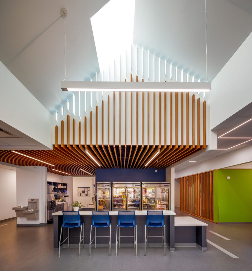 Modern cafeteria interior with a row of blue chairs at a counter, wooden ceiling slats, refrigerators in the back, and a skylight overhead.