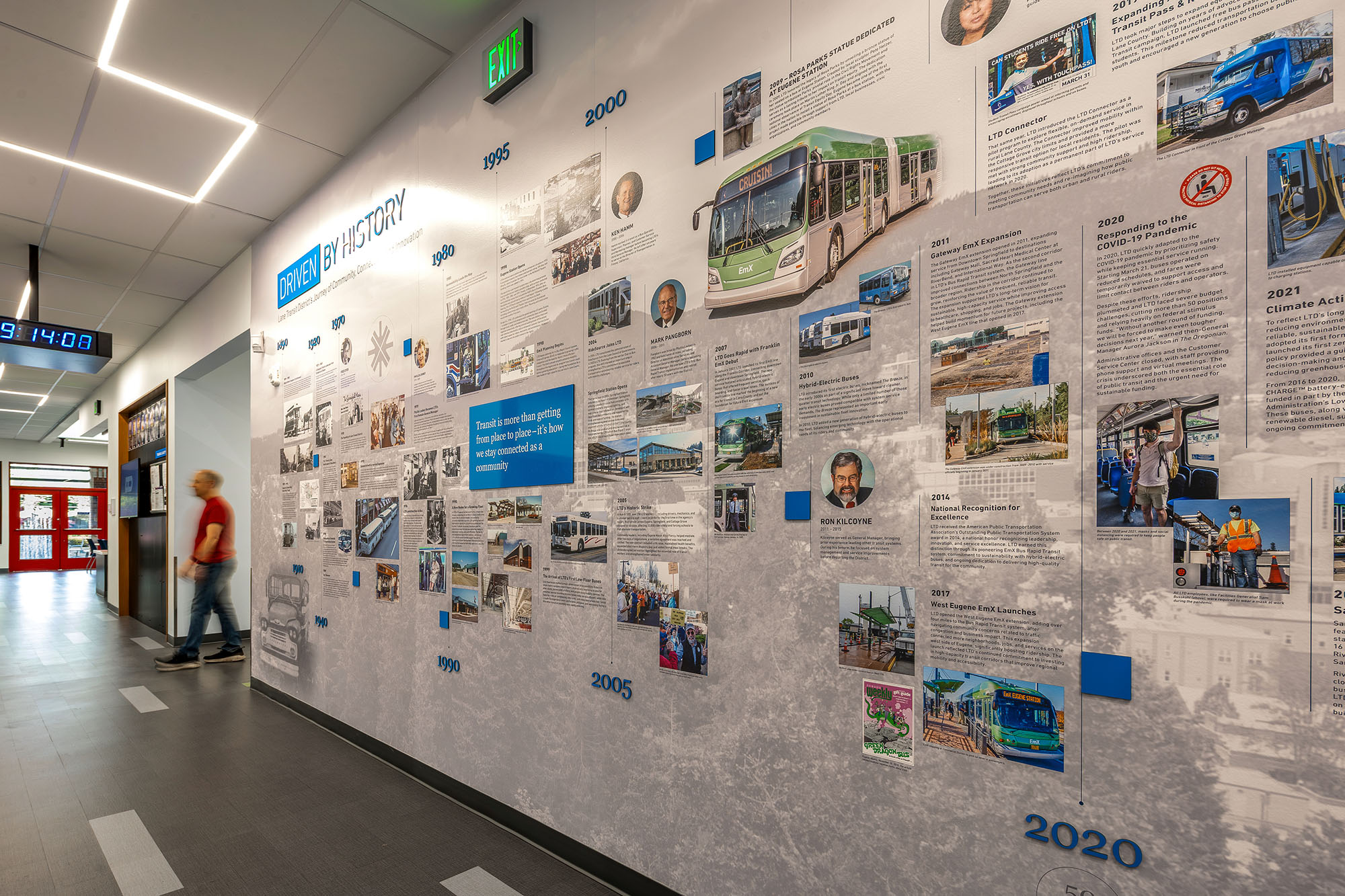 A person walks past a large timeline display on a hallway wall, featuring photos and text about transit history from 1985 to 2021.