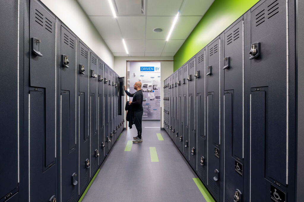 A person stands in a modern locker room with rows of dark lockers and a brightly lit ceiling.