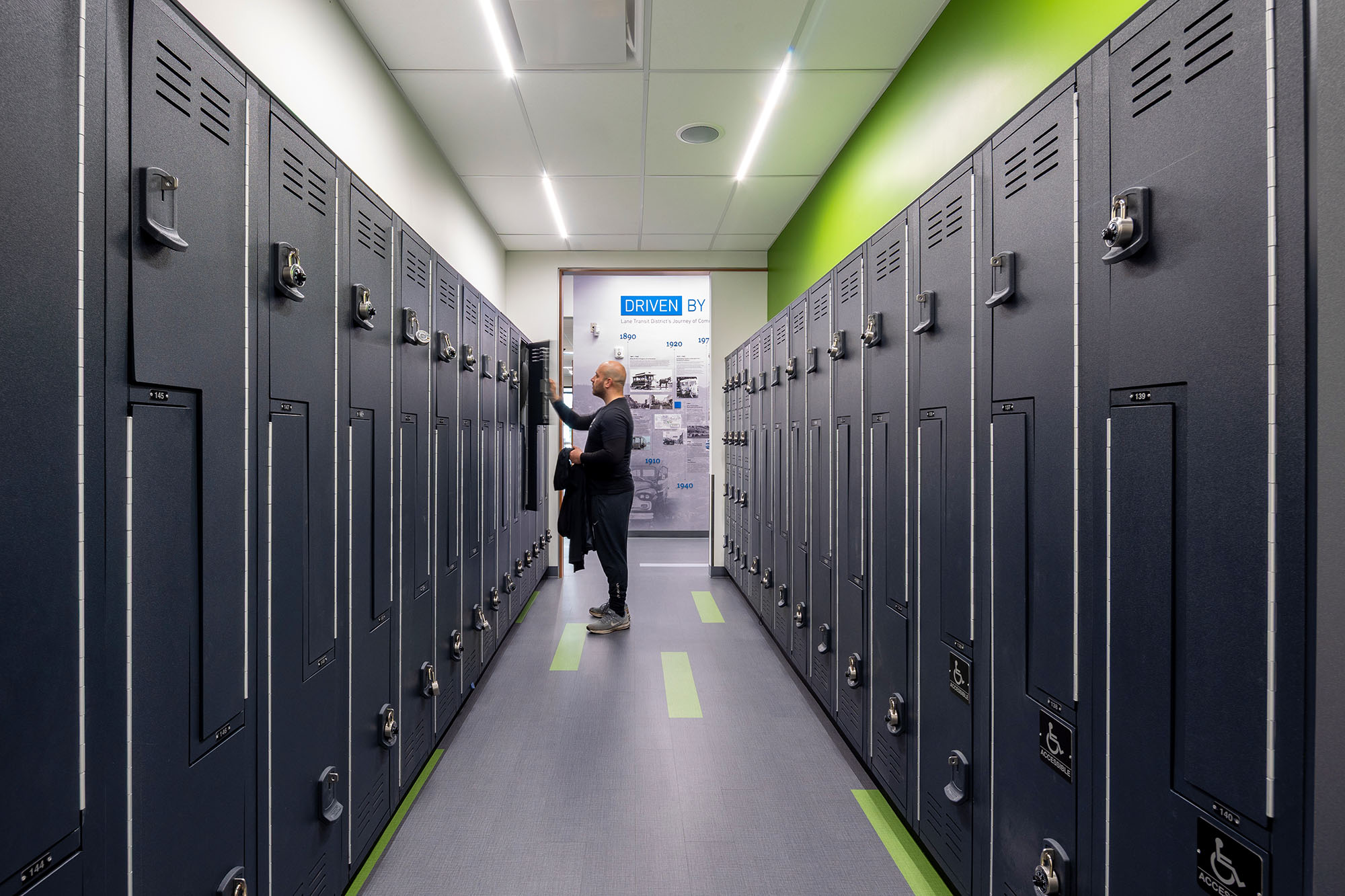 A person stands in a modern locker room with rows of dark lockers and a brightly lit ceiling.
