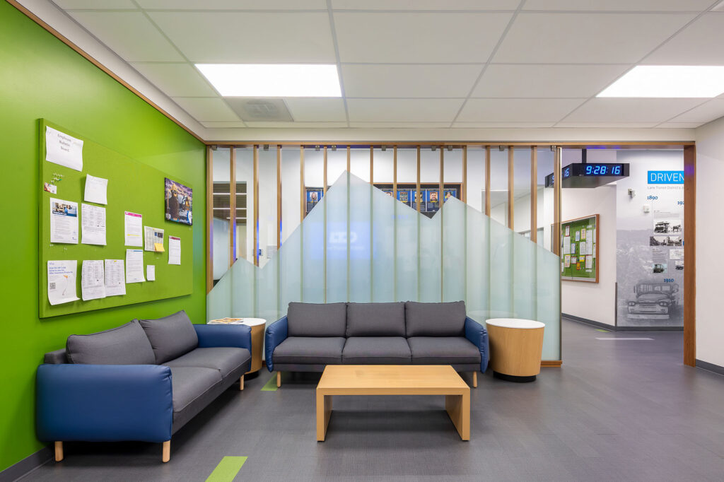 Modern waiting area with two blue and gray sofas, a wooden coffee table, green accent wall with bulletin board, and decorative glass partition. Hallway visible in the background.
