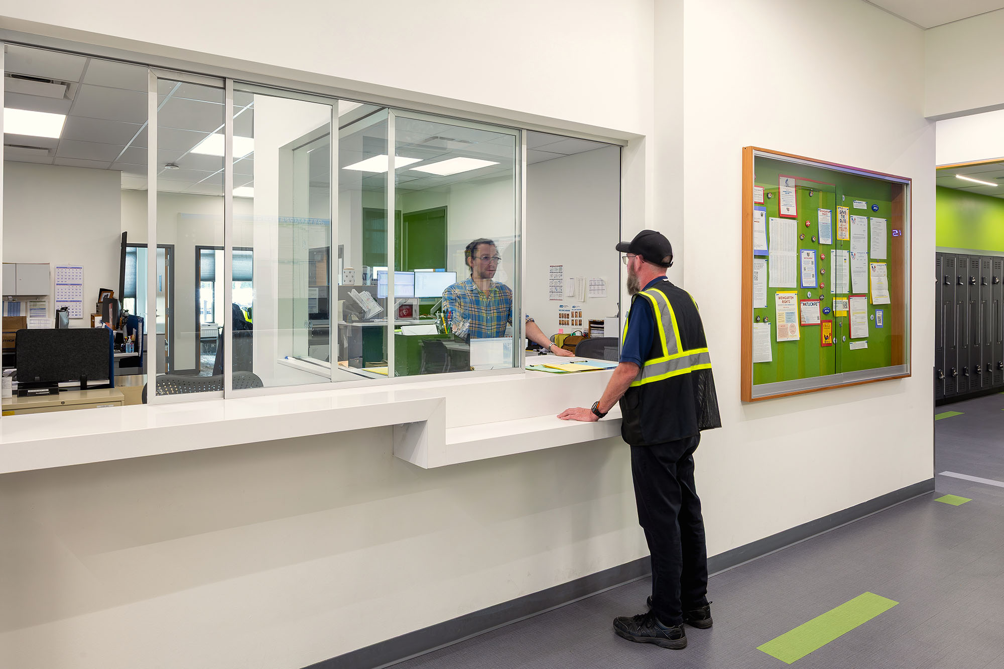 A person in a safety vest speaks to a staff member behind a glass window at a reception or service counter in a modern office or facility.