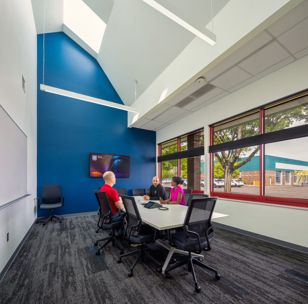 Three people sit at a conference table in a modern meeting room with large windows, a blue accent wall, and a wall-mounted screen.