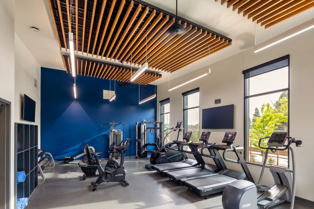 Modern gym with treadmills, stationary bikes, weight machines, TVs, blue accent wall, large windows, and wooden slat ceiling.