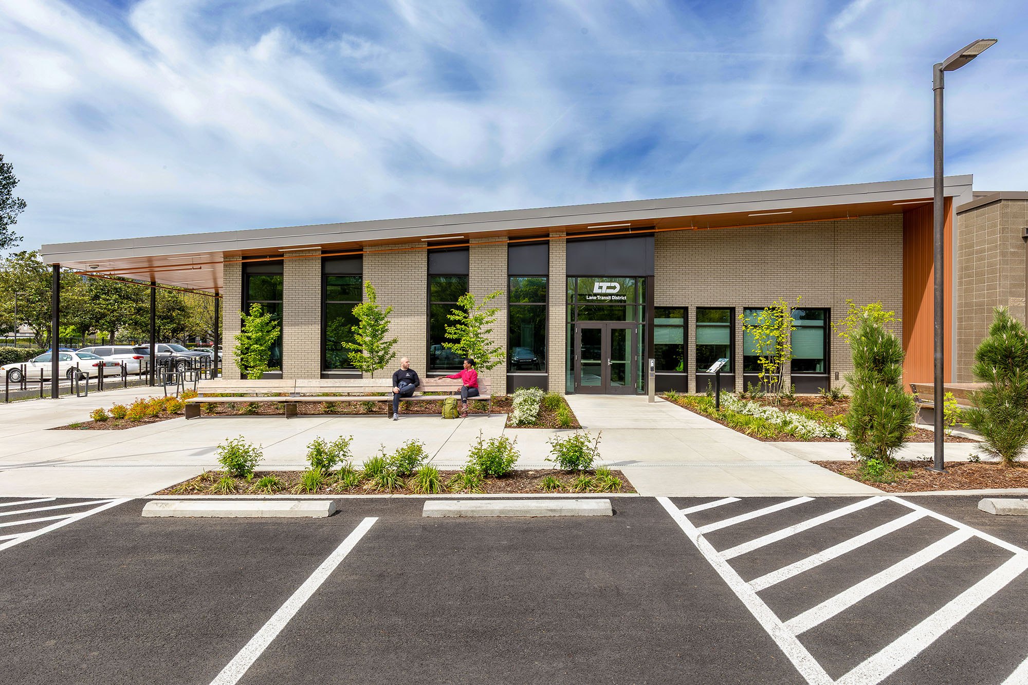 Modern building with large windows, landscaped entrance, benches, and two people sitting outside; empty parking lot in the foreground.