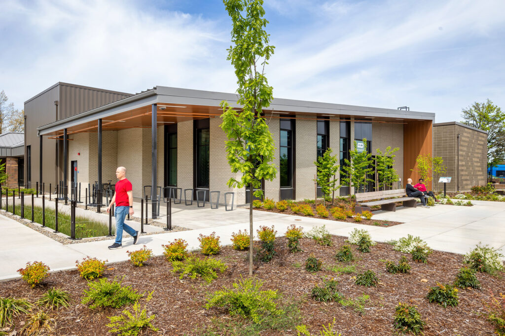 A modern, single-story building with large windows and a covered entrance; landscaped grounds and a few people outside on a sunny day.