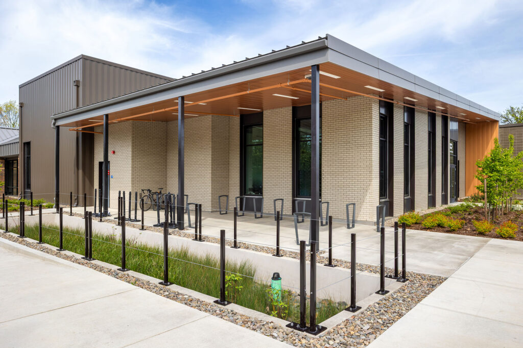 Modern building with large windows, a covered walkway, bike racks, and landscaped surroundings under a partly cloudy sky.