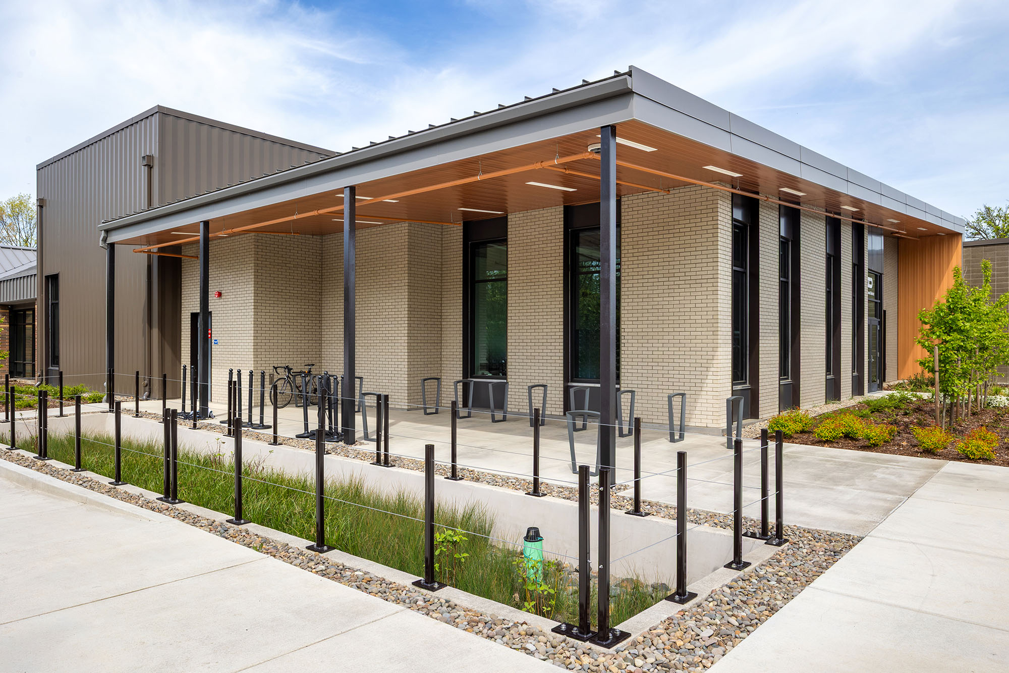 Modern building with large windows, a covered walkway, bike racks, and landscaped surroundings under a partly cloudy sky.