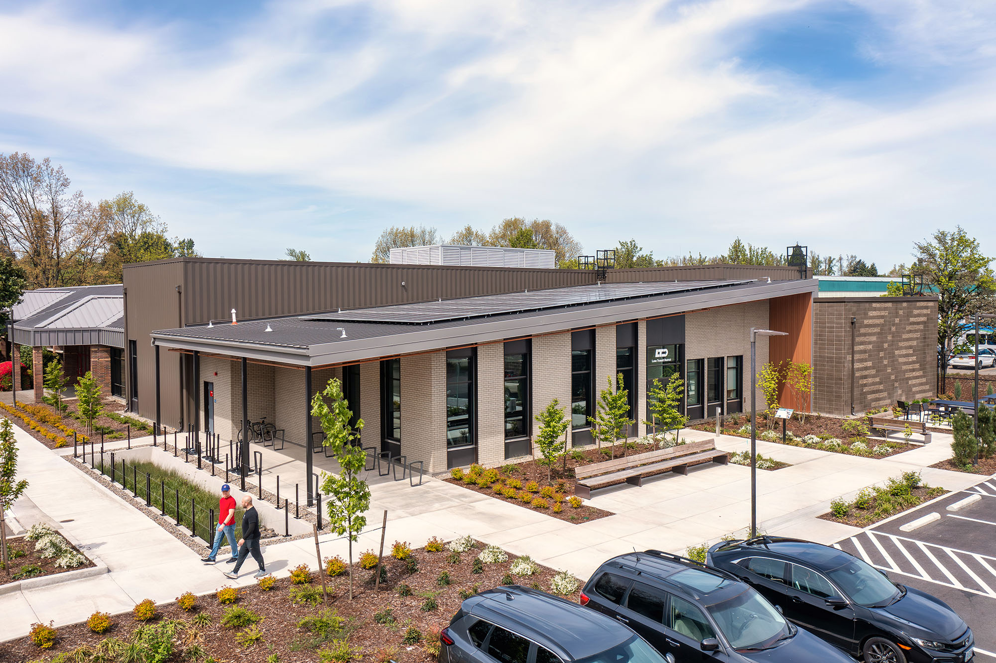 Modern, single-story building with large windows, flat roofs, and landscaped surroundings; two people walk on a sidewalk near parked cars under a partly cloudy sky.