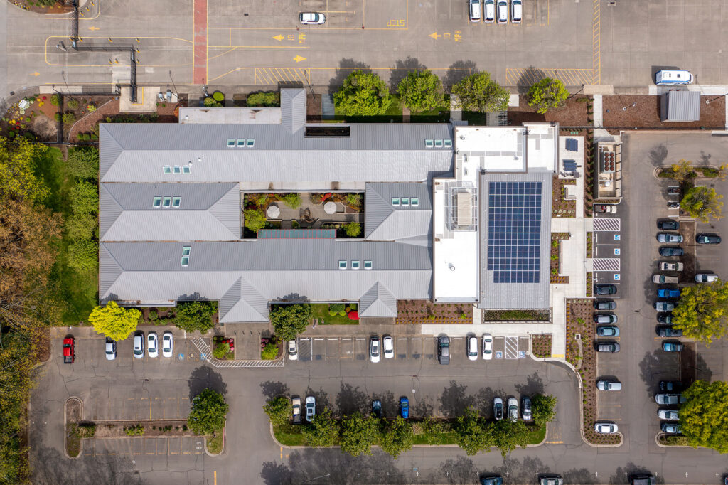 Aerial view of a large building with solar panels on the roof, surrounded by parking lots with cars and landscaped green areas.