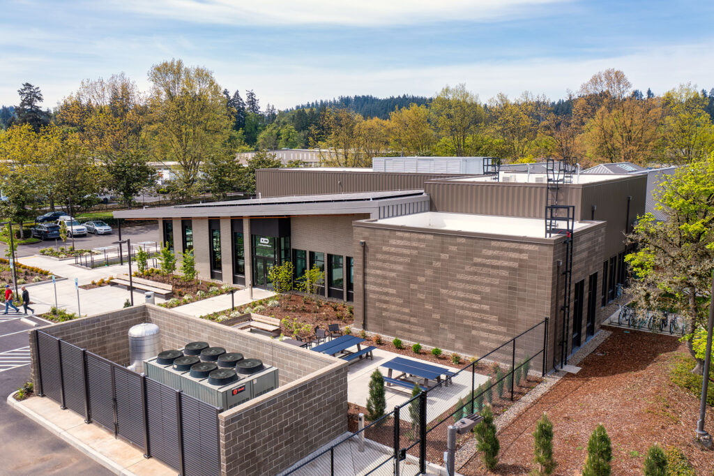 A modern, single-story commercial or institutional building with large windows, outdoor seating, landscaping, and a fenced mechanical area, surrounded by trees and parking spaces.