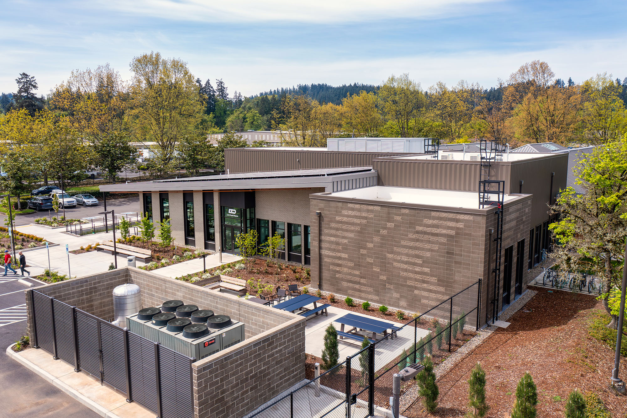 A modern, single-story commercial or institutional building with large windows, outdoor seating, landscaping, and a fenced mechanical area, surrounded by trees and parking spaces.