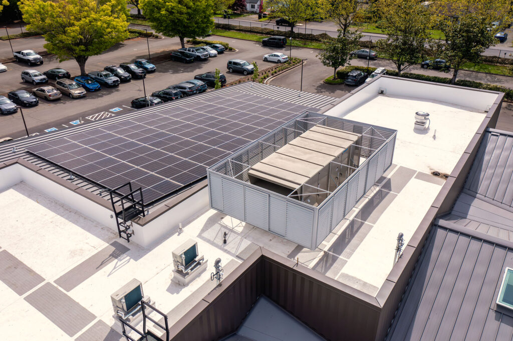 Aerial view of a flat building roof with solar panels, HVAC units, and a fenced mechanical enclosure, next to a parking lot with cars and green trees.